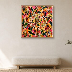 A framed square colorful abstract artwork hangs centered on a plain wall above a minimalist neutral bench and a small wooden side table with a vase.