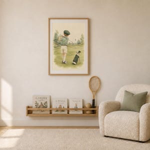 May include: A framed watercolor print depicting a child in a green cap and shirt playing golf on a grassy course with a golf bag nearby. The artwork is displayed on a wall above a wooden shelf holding three books and a vintage wooden tennis racket.
