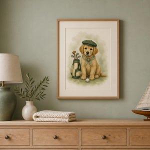 May include: A framed wall art print featuring a golden retriever puppy wearing a green cap and a neckerchief. The puppy sits beside a golf bag filled with clubs and a single golf ball on the ground. The illustration uses soft, muted tones.