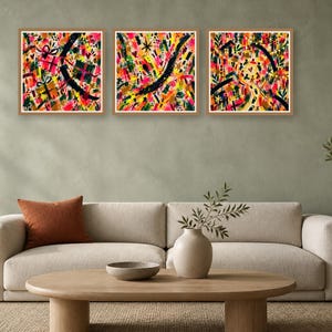 Living room interior featuring a beige sofa, wooden coffee table, and three square colorful abstract artworks hung on a sage green wall, showing different prints.