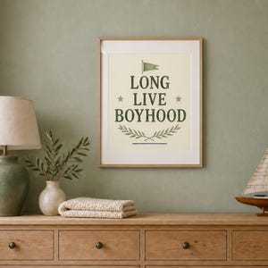 Puede incluir: Una l&aacute;mina de arte mural enmarcada con fondo color crema y el texto verde oscuro "LONG LIVE BOYHOOD" centrado. El dise&ntilde;o incluye un peque&ntilde;o icono de bandera verde sobre el texto, dos estrellas a cada lado y un motivo de rama con hojas debajo.