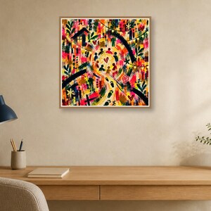 A framed square abstract artwork hanging on a beige wall above a wooden desk with a lamp, pencil cup, and notebook.