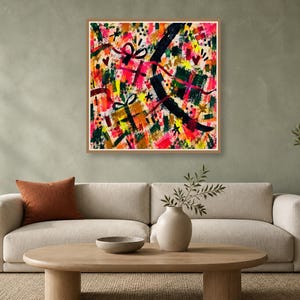 A wood-framed square abstract art print with vibrant brushstrokes hangs above a neutral sofa on a green wall.
