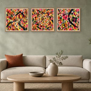 A living room interior featuring a beige sofa with a rust pillow, a wooden coffee table, and three framed abstract square art prints hanging on a grey wall.