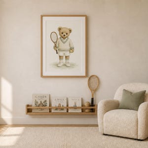 May include: A framed wall art print featuring a light brown teddy bear dressed in a white tennis sweater and shorts, holding a tennis racket. The bear stands on a patch of green grass against a plain, light-colored background within a wooden frame.