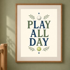 May include: A framed wall art print featuring the text PLAY ALL DAY in alternating dark blue and olive green serif letters. The design includes a white golf ball at the top, a yellow-green tennis ball at the bottom, and small leaf sprigs.