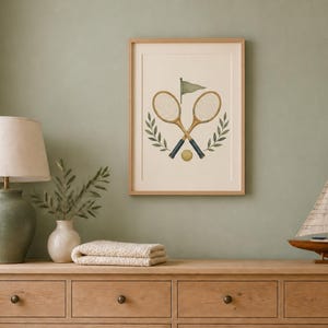 May include: A framed wall art print featuring a centered illustration of two crossed vintage wooden tennis rackets, a small yellow tennis ball, a green flag, and two symmetrical green leafy branches on a cream-colored background.