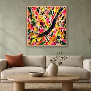 A square wood-framed abstract art print featuring bright colorful strokes hangs over a beige sofa in a room with a green wall.