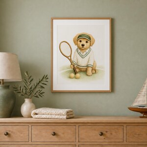 May include: A framed wall art print featuring a golden retriever puppy wearing a white tennis sweater with green trim and a matching green visor. The puppy holds a wooden tennis racket in one paw, with two yellow tennis balls resting on the ground.