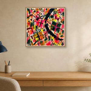 A square abstract art print in a thin frame hangs on a neutral wall above a wooden desk with a lamp and notebook.