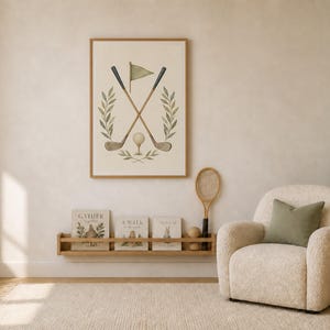 May include: A rectangular framed wall art print featuring a vintage-style illustration of two crossed golf clubs, a golf ball, a small flag, and green leafy branches on a cream background, displayed in a room with a cream armchair and shelf.
