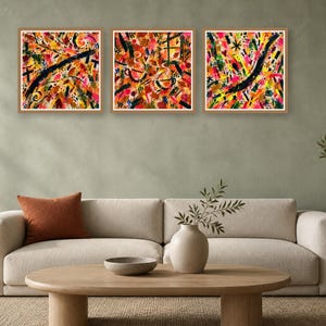 Living room interior featuring a beige sofa with an orange pillow and three square abstract colorful wall art prints hanging above it.