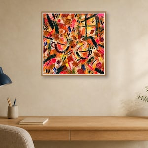 A square wood-framed abstract painting hangs above a wooden desk with a blue desk lamp, notebook, and potted plant.