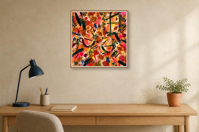 A square wood-framed abstract painting hangs above a wooden desk with a blue desk lamp, notebook, and potted plant.