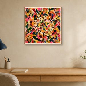 A framed square abstract art piece is mounted on a beige wall above a wooden desk setup featuring a blue lamp, notebook, and potted plant.