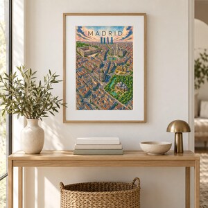 K&ouml;nnte beinhalten: Ein gerahmter Kunstdruck mit einer Luftaufnahme-Illustration von Madrid und dem Schriftzug MADRID am oberen Rand. Das Bild zeigt ein dichtes Stadtbild mit orangefarbenen D&auml;chern, gr&uuml;nen Parkanlagen und einem bew&ouml;lkten blauen Himmel.
