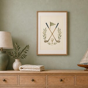 May include: A framed wall art print featuring a watercolor illustration of two crossed golf clubs, a golf ball, a flag, and surrounding olive branches. The artwork is displayed in a light wood frame with a cream-colored background and neutral tones.
