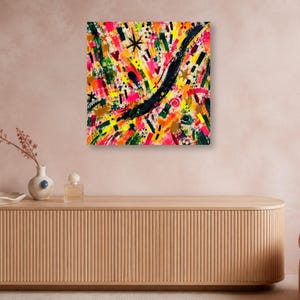 A square colorful abstract art print is mounted on a pinkish wall above a long wooden credenza with vases and a perfume bottle.