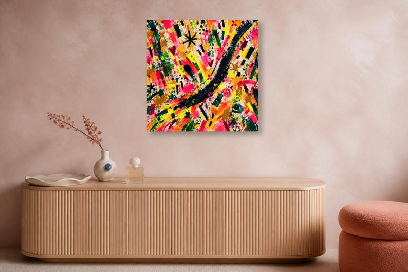 A square colorful abstract art print is mounted on a pinkish wall above a long wooden credenza with vases and a perfume bottle.