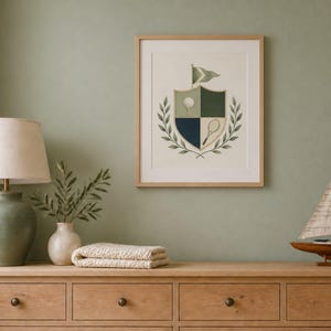 Puede incluir: Una l&aacute;mina de arte enmarcada que muestra un escudo con una pelota de golf, una raqueta de tenis y una bandera, rodeado de ramas de laurel. El dise&ntilde;o utiliza tonos verdes, crema y azul oscuro, presentado en un marco de madera clara.