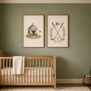 May include: Two framed wall art prints featuring golf-themed illustrations. One print shows a small golf clubhouse with a flag, and the other displays two crossed golf clubs, a golf ball, and a flag, all framed in light wood on a sage green wall.