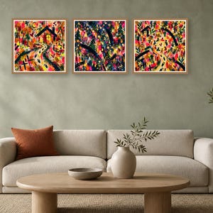 Three framed square abstract art prints with colorful gestural strokes hang above a modern beige sofa in a minimalist living room.