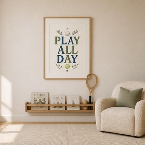 May include: A framed wall art print featuring the text PLAY ALL DAY in dark green and blue letters, accented with illustrations of a white golf ball, a yellow tennis ball, and green leaves, displayed in a light wood frame on a neutral wall.