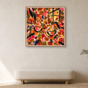 A square wood-framed abstract print with warm autumn tones hangs centered on a plain wall above a low beige upholstered bench.