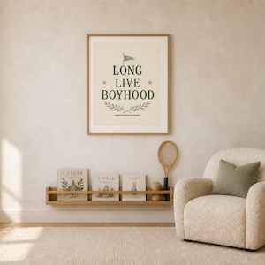 Puede incluir: Un cuadro de pared con el texto LONG LIVE BOYHOOD en letras de color verde oscuro, acentuado por una peque&ntilde;a bandera, dos estrellas y un dise&ntilde;o de rama de laurel. El cuadro se muestra sobre un estante de madera con tres libros y una raqueta.