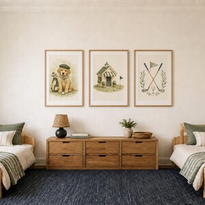 May include: Three framed wall art prints featuring golf-themed illustrations: a golden retriever puppy wearing a cap, a small clubhouse building, and crossed golf clubs with a flag. The prints are displayed in light wood frames on a white wall.