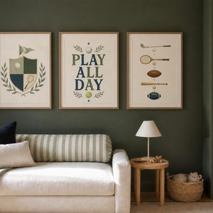 May include: Three framed wall art prints featuring sports themes. The left print shows a shield with a golf flag, tennis racket, and laurel leaves. The center print displays the text "PLAY ALL DAY" with sports balls. The right print shows various sports equipment.