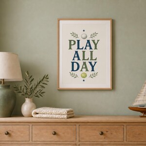 May include: A framed wall art print featuring the text PLAY ALL DAY in dark blue and olive green lettering, accented with illustrations of a white golf ball, a yellow-green tennis ball, small dark blue stars, and green leafy sprig motifs.