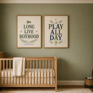 Puede incluir: Dos l&aacute;minas decorativas enmarcadas con bordes claros colgadas sobre una cuna de madera. La l&aacute;mina de la izquierda presenta el texto LONG LIVE BOYHOOD con una bandera y motivos de hojas. La de la derecha muestra PLAY ALL DAY con una pelota de golf.
