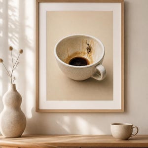 A framed art print of a coffee cup hangs above a wooden credenza with a ceramic vase and smaller cup.