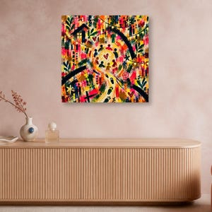 A square abstract artwork hung on a pink wall above a long slatted wooden sideboard featuring a vase with branches and perfume bottles.