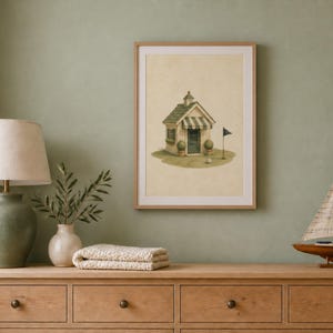May include: A framed wall art print featuring a watercolor illustration of a small golf clubhouse with a striped awning, a green roof, and a golf flag on a putting green. The artwork is displayed in a light wood frame on a sage green wall.
