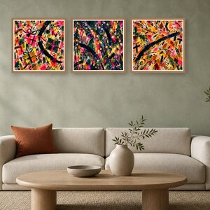 A neutral living room features a beige sofa with an orange pillow, a wood coffee table, and three matching square abstract art prints in light wood frames hung on a green wall.