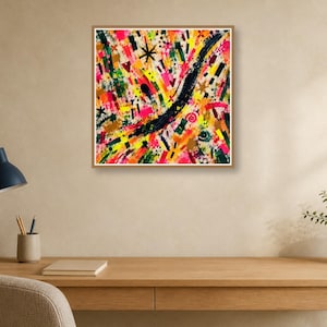 A framed abstract print hangs above a wooden desk with a black lamp, notebook, and pen against a warm beige wall.