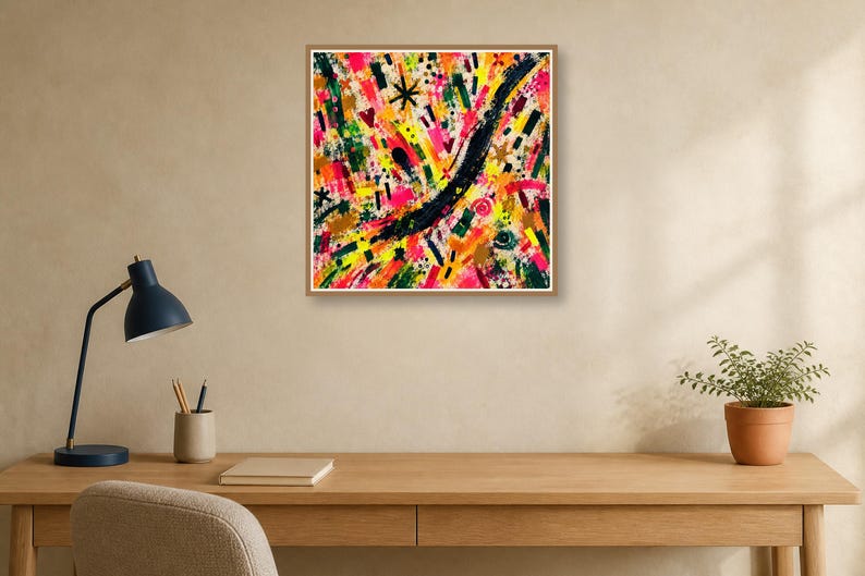 A framed abstract print hangs above a wooden desk with a black lamp, notebook, and pen against a warm beige wall.