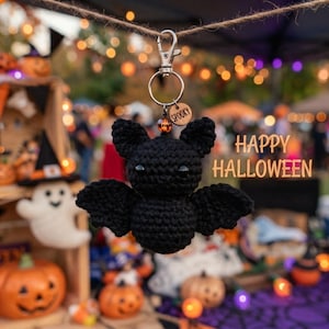 Könnte beinhalten: Ein schwarzer, gehäkelter Fledermaus-Schlüsselanhänger mit einem silbernen Karabinerhaken, einem kleinen runden Holzanhänger mit der Gravur SPOOKY und einer bernsteinfarbenen Perle. Die Fledermaus hat zwei spitze Ohren und zwei breite Flügel.