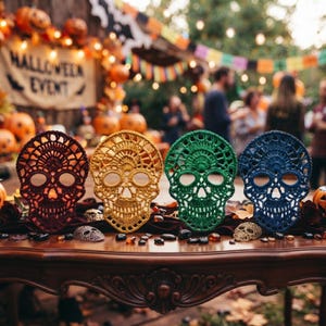 Puede incluir: Cuatro decoraciones de calaveras de ganchillo en rojo oscuro, amarillo brillante, verde bosque y azul marino. Cada calavera presenta intrincados patrones de encaje calado con grandes cuencas oculares y una cavidad nasal detallada sobre una mesa.