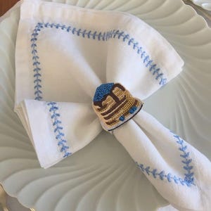 May include: A white cloth napkin with a light blue embroidered border, held by a beaded napkin ring shaped like a stack of pancakes with blue berries on top. The napkin rests on a white scalloped plate with a silver utensil visible nearby.