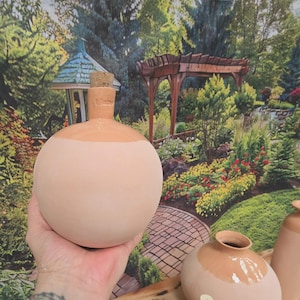 May include: A spherical terracotta vessel with a smooth, matte finish on the lower portion and a glossy, glazed finish on the upper neck area. The vessel features a narrow opening sealed with a natural cork stopper and the word Vintner embossed.