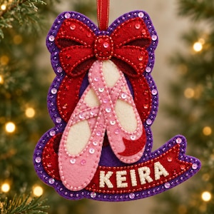 May include: A felt Christmas ornament featuring a pair of light pink ballet slippers centered below a large red bow. The design includes a purple backing, red ribbon accents, and sequins throughout. A red banner at the bottom displays the name KEIRA.