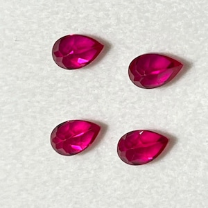 Puede incluir: Cuatro piedras preciosas sueltas en forma de pera con un color magenta rojizo intenso y vibrante. Cada piedra presenta una superficie facetada que refleja la luz, creando un aspecto brillante y centelleante. Las gemas están dispuestas en cuadrícula.