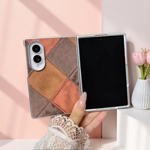 May include: A foldable smartphone case featuring a patchwork design of suede-like squares in shades of brown, tan, and burnt orange. The case includes a cutout for a dual-lens camera system and a black screen area, showcasing a textured finish.