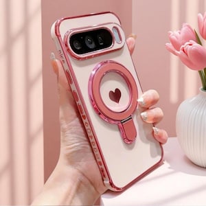 May include: A light pink smartphone case with a metallic pink frame and camera surround. The back features a circular magnetic ring with a small heart icon in the center. The side edge is printed with the word nice and small heart symbols.