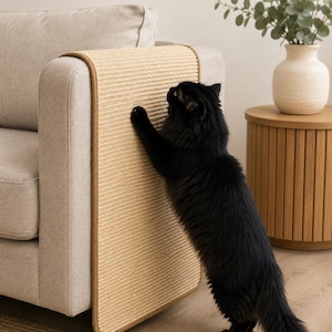 Puede incluir: Una alfombrilla rascadora rectangular de sisal beige con un borde de tela marrón, colocada sobre el brazo de un sofá tapizado en gris claro. La alfombrilla presenta una superficie tejida texturizada y está colocada verticalmente sobre el brazo.