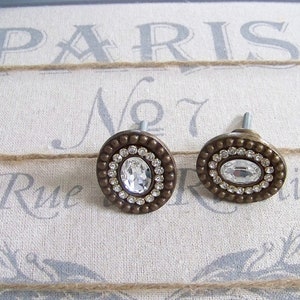 May include: Two oval-shaped cabinet knobs with a bronze finish. Each knob features a large, clear crystal rhinestone surrounded by smaller rhinestones.