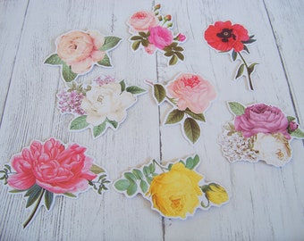 May include: Assortment of eight paper flower embellishments with various colors and designs, including pink, white, yellow, red, and purple. The flowers are cut out and have a white background.